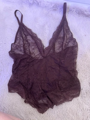 Mörklila spetsväst med v-ringning - Säljer en mörklila väst i transparent spets med blommigt mönster. Västen har smala axelband och djup v-ringning både fram och bak. Materialet är lätt och luftigt, perfekt för att styla lager på lager eller som statement-plagg.