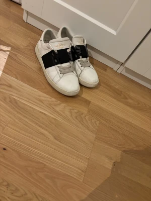 Valentino Garavani Open sneakers vit/svart - Säljer ett par Valentino Garavani Open sneakers i storlek 42. Skorna är vita med en bred svart rem över mitten och har grå snörning. Tillverkade i skinn med rund tå och platt sula. Finns tecken på användning så bara fråga. 