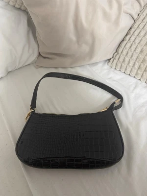 Svart axelväska i croco-mönster - Snygg svart axelväska med croco-mönstrad yta och guldfärgade detaljer. Väskan har en kort rem och stängs med dragkedja. Perfekt storlek för det viktigaste och ger en trendig touch till din outfit.