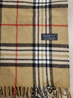 Burberrys Halsduk Beige Vintage 90s - Klassisk halsduk från Burberrys med det ikoniska rutiga mönstret i beige, svart, vitt och rött. Halsduken har fransar i ändarna och är mjuk och varm, perfekt för kyliga dagar.