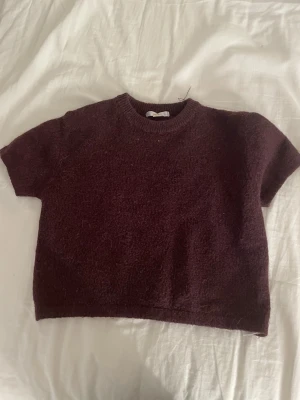 Vinröd stickad croppad tröja - Säljer en vinröd stickad tröja med korta ärmar och croppad passform. Tröjan har rund halsringning och är gjord i ett mjukt stickat material som känns skönt mot huden. Perfekt för lager-på-lager och att styla med höga jeans.