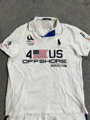 Vit RLX Polo Offshore Racing skjorta - Säljer en vit kortärmad skjorta från RLX Polo Ralph Lauren med tryck och broderade detaljer. Skjortan har klassisk krage, blå insida vid halsen och stora tryck med amerikansk flagga och texten '4 US Offshore Racing' både fram och bak. Perfekt för dig som gillar sportig stil. 