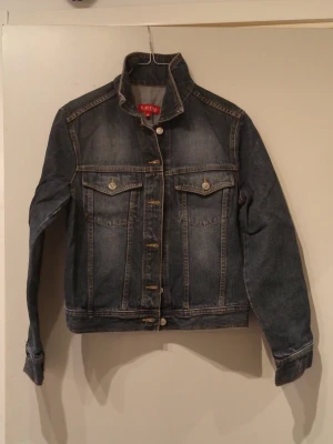 Broderad jeansjacka från L.O.G.G. - Snygg mörkblå jeansjacka från L.O.G.G. i klassisk modell med knappar framtill och två bröstfickor. Baksidan har färgglad broderad design med ansikte, fjärilar och blommor som ger jackan en unik vibe. Perfekt för dig som vill sticka ut med din stil.