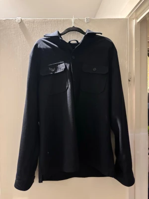 Svart overshirt från Riley - Säljer en mörkblå overshirt från Riley i slim fit-modell. Jackan har två stora bröstfickor med knappar, klassisk krage och knäppning framtill. Materialet känns mjukt och passar perfekt för lager-på-lager. Enkel och stilren design som funkar till det mesta.