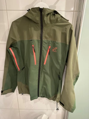 Grön vindjacka från Revolution Race - Säljer en grön vindjacka från Revolutionrace i modellen silence proshell 3L i färgen forest night. På hemsidan är denna storlek slutsåld. Jackan har huva och flera fickor med korall dragkedjor. Jackan har justerbara ärmslut med kardborre och dragsko nedtill. Perfekt för utomhusaktiviteter och har en cool tvåtonad grön design. Går bra att åka skidor i. Den är lite sliten invändigt vilket syns på bilderna men fortfarande snygg utvändigt 