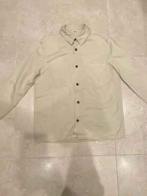 Beige overshirt jacka med fickor från ARKET - Snygg beige overshirt jacka i bomull med klassisk krage och svarta knappar framtill. Två stora bröstfickor ger en clean och stilren look. Perfekt att slänga på över en hoodie eller t-shirt för en chill vibe.