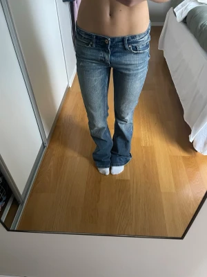 Blå bootcut jeans låg midja - Säljer ett par blå jeans med snygg bootcut och låg midja. Jeansen har klassisk femficksdesign, bälteshällor och dragkedja med knapp. Materialet är jeans i bomull och passformen är normal med lätt utsvängda ben. Perfekt för dig som gillar retrovibbar och avslappnad stil.