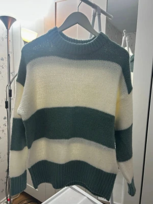 Randig stickad tröja i grönt och vitt - Säljer en mysig stickad tröja med breda ränder i mörkgrönt och vitt. Tröjan har rund hals, ribbade muddar och långärmad passform. Perfekt för dig som gillar en chill och avslappnad stil med lite oversized känsla.