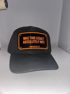 Svart truckerkeps från John Hatter & Co - Säljer en svart truckerkeps från John Hatter & Co med mesh på sidorna och bak. Framtill sitter en metallskylt med orange text: 'WAS THIS LEGAL? ABSOLUTELY NOT!'. Kepsen har böjd skärm och justerbar snapback. Perfekt statement piece för dig som gillar att sticka ut. Lådan ingår!