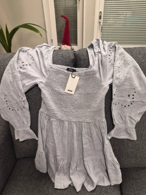 Bik Bok DW Air grå klänning, storlek L - Supersöt ljusgrå klänning från Bik Bok med smockad överdel och volang i kjolen. Klänningen har långa ärmar med broderade detaljer och elastiska muddar. Materialet känns mjukt och luftigt, perfekt för vår och sommar. Fyrkantig urringning ger en trendig vibe.
