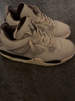 Nike Air Jordan 4 beige sneakers - Säljer ett par Nike Air Jordan 4 sneakers i beige med svarta detaljer och meshpaneler på sidorna. Skorna har snörning, platt sula och rund tå. Ikonisk Jumpman-logga på plösen och Nike Air-märke på hälen. Perfekt för dig som gillar streetwear och snygga sneakers.