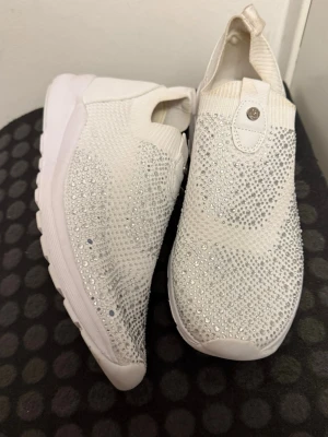 Vita sneakers med glittriga detaljer - Säljer ett par vita sneakers med glittriga stenar över hela ovandelen. Skorna har en stickad ovandel i syntetmaterial och en tjock, vit platt sula. De är slip-on och har en modern, sportig look med extra bling.