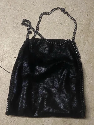 Stella McCartney liknande väska  - Säljer en svart axelväska med glansig finish och snygga kedjedetaljer längs kanterna och som axelrem. Väskan har en mjuk, slouchy form och passar perfekt för dig som gillar en cool och stilren look. Materialet känns som syntet och ger en modern vibe.