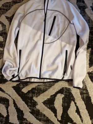 Vit Fleecejacka från Bluewear L - Säljer en vit jacka från Bluewear i storlek L. Jackan har svarta detaljer, tre dragkedjeförsedda fickor och snygga sömmar som ger en sportig vibe. Perfekt för dig som gillar stilren och funktionell design.