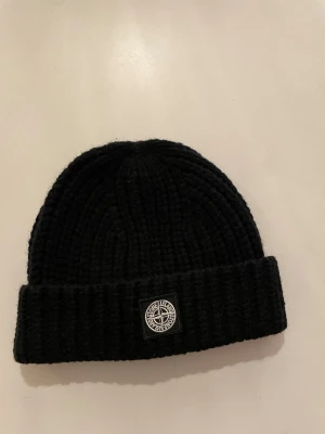 Svart mössa från Stone Island - Säljer en svart ribbstickad mössa från Stone Island med uppvikt kant och klassisk logotyp framtill. Mössan är tillverkad i mjuk ull och har en enkel, stilren design som passar perfekt för kalla dagar.