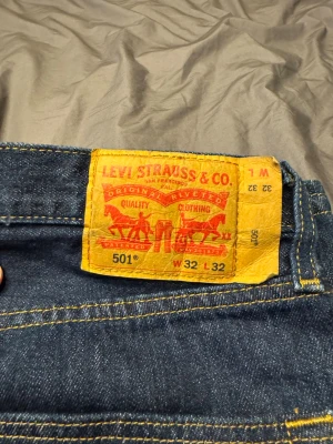 Levi's 501 blå jeans W32 L32 - Klassiska Levi's 501 jeans i mörkblå denim med raka ben och gul kontrastsöm. Modellen har fem fickor och knappgylf. Perfekta för dig som gillar tidlös stil och vill ha ett par riktigt schyssta jeans i bomull.