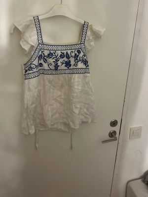 Vit blus med blå broderi och volang - Säljer en vit blus med blå broderade blommor och rutmönster upptill. Blusen har volangdetaljer vid axlarna och knytband i sidorna. Den är ärmlös med fyrkantig halsringning och tillverkad i ett lätt, luftigt material som passar perfekt till varma dagar.Pris kan diskuteras och tvätt ingår     