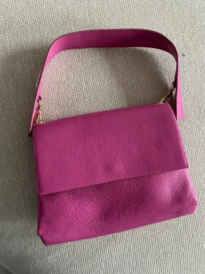 Rosa axelväska i skinn - Säljer en snygg rosa axelväska i skinn med stilren design och guldiga detaljer. Väskan har ett brett axelband och praktiskt magnetlås. Insidan har flera fack för enkel organisering av dina grejer. Tillkommer även ett axelremsband.