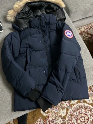 Navy blue Canada Goose med päls - Säljer en Navy Blue Canada Goose med ikonisk patch på ärmen och fluffig päls på huvan. Jackan har dragkedja, långa ärmar och flera fickor. Insidan är fodrad med svart, glansigt material. Perfekt för kalla vinterdagar. Köpte den men den passar inte mig tyvärr så väljer att sälja den, priset kan diskuteras vid snabb försäljning!