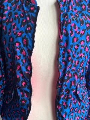 Rosa och leopardmönstrad vindjacka Cars Jeans - Säljer en vändbar vindjacka från Cars Jeans med ena sidan i stark rosa och broderad blå tiger, andra sidan i blått leopardmönster med rosa och svart detaljer. Jackan har dragkedja och är tillverkad i polyester. Perfekt för dig som gillar att sticka ut.