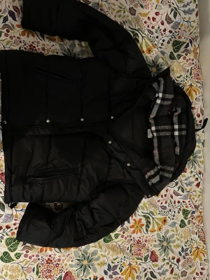 Svart pufferjacka från Burberry - Svart pufferjacka från Burberry med avtagbara ärmar så den kan användas som väst. Jackan har dragkedja, tryckknappar och två fickor med dragkedja. Huva med klassiskt rutigt Burberry-mönster i grått, vitt och rött. Perfekt för kalla dagar och riktigt snygg streetstil.