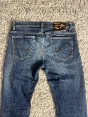 Blå jeans från Jacob Cohen - Säljer ett par klassiska blå jeans från Jacob Cohen med snygg tvätt och kontrastsömmar. Modellen har fem fickor, normal passform och raka ben. Märkespatch i brunt på linningen bak. Perfekta till sneakers eller boots.