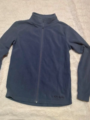 Blågrå fleecejacka från Stormberg - Mjuk och skön blågrå fleecejacka från Stormberg med hel dragkedja framtill och hög krage. Perfekt för kyliga dagar och enkel att slänga på sig över en t-shirt. Diskret logga broderad längst ner på framsidan. Långärmad och normal passform.