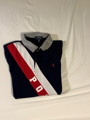Mörkblå piké från Polo Ralph Lauren - Säljer en mörkblå pikétröja från Polo Ralph Lauren med grå krage och ärmslut. Framsidan har ett diagonalt rött och vitt band med texten 'POLO' och en liten röd logga på bröstet. Klassisk och sportig design, perfekt för dig som gillar stilrena detaljer.