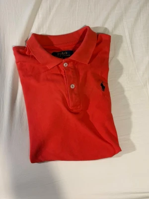 Röd pikétröja från Polo Ralph Lauren - Säljer en klassisk röd pikétröja från Polo Ralph Lauren med korta ärmar och krage. Tröjan har två knappar framtill och den ikoniska broderade loggan i marinblått på bröstet. Perfekt för dig som gillar stilrena och sportiga plagg. S passar M 