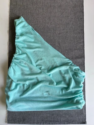 Turkos one-shoulder topp, storlek L - Säljer en snygg turkos topp i storlek L. Jag är normalt en S och den sitter bra på mig 😊 Rynkade detaljer längs sidan och tillverkad i ett mjukt material. Använd en gång! - Köper du innan kl 16:00 kan jag posta samma dag!