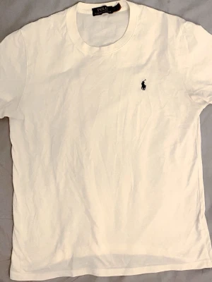 Vit t-shirt från Polo Ralph Lauren - Vit t-shirt från Polo Ralph Lauren med klassisk rund hals och korta ärmar. På bröstet finns den ikoniska broderade polospelaren i svart. T-shirten är gjord i mjuk bomull och har en clean, stilren look som funkar till allt.