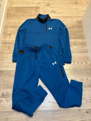 Under Armour tracksuit - Snyggt blått träningsset från Under Armour med matchande tröja och byxor. Tröjan har svart krage, half zip och vit logga på bröstet samt tryck på ärmen. Byxorna har dragsko i midjan, svarta detaljer vid fickorna och vit logga på benet. Perfekt för gym eller sport.