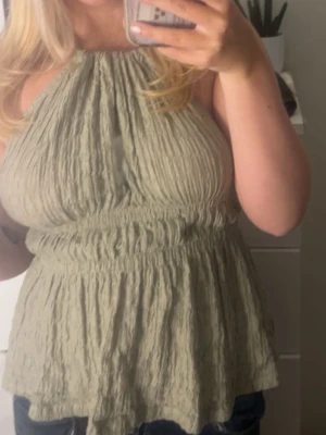 Grön ärmlös blus med rynk - Säljer en ljusgrön blus med rynkad struktur och halterneck. Toppen är ärmlös och har en lös passform med smock i midjan för en snygg siluett. Perfekt för varma dagar och enkel att matcha med jeans eller shorts. Blusen är helt öppen i ryggen, skriv för bilder