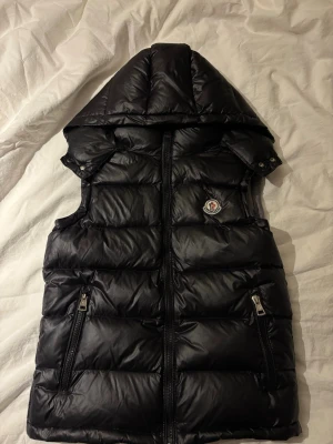 Moncler väst - Hallö! Säljer en svart dunväst från Moncler med vadderad huva och två sidofickor med dragkedja. Västen har quiltad design, Moncler-logga på bröstet och en snygg, glansig finish. Perfekt att slänga över en hoodie eller t-shirt för extra värme. Fet och timeless väst.💫 Storlek L men sitter mer som en liten M/ S💵💵