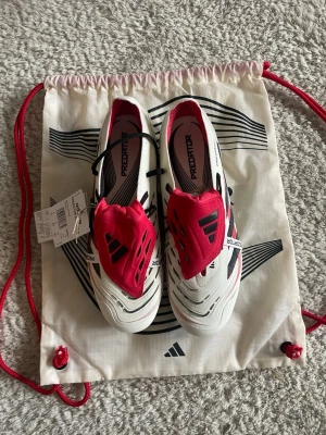 Adidas Predator Elite FT S fotbollsskor Storlek 42 - Adidas Predator Elite FT S fotbollsskor i vitt med röda och svarta detaljer. Skorna har snörning och en täckt plös med Predator-logga. Sulan har dobbar för optimalt grepp på planen och HYBRIDTOUCH-material för bra känsla. Perfekt för dig som vill ha max kontroll och stil på fotbollsplanen. Kommer med påse och extra metallskruvdobbar. Ordinarie pris:3099kr