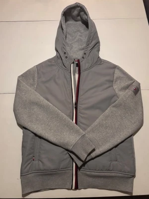 Grå Tommy Hilfiger cardigan - Snygg grå cardigan från Tommy Hilfiger med huva och dragkedja framtill. Jackan har mjuka ärmar och detaljer i rött, vitt och marinblått längs dragkedjan. Perfekt för dig som gillar stilrena och sportiga plagg med ikonisk logga på ärmen och ryggen. Hör av dig om du skulle ha någon fråga!