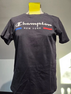 Svart Champion t-shirt New York - Svart t-shirt från Champion med trycket 'Champion New York' på bröstet och detaljer i blått och rött. Klassisk passform och rund hals. Perfekt för dig som gillar streetwear och vill ha en enkel men snygg look.