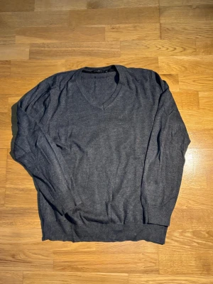 Mörkgrå stickad tröja från DKNY - Snygg mörkgrå stickad tröja från DKNY med v-ringad hals och långa ärmar. Tröjan har ribbade muddar vid ärmslut och nederkant, vilket ger en schysst passform. Perfekt för dig som gillar stilrena och enkla plagg med lite edge.