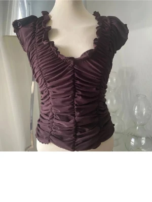  omlott topp med volang - Säljer en brun  topp med omlott-effekt och volangdetaljer vid halsringningen. Toppen har korta ärmar och är rynkad framtill för en snygg siluett. Materialet känns mjukt och stretchigt, perfekt för att lyfta din outfit.