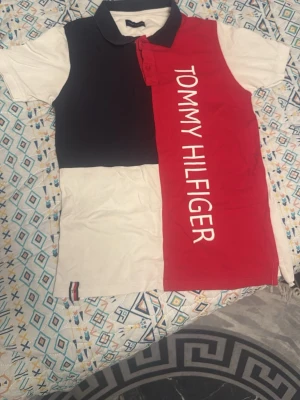 Färgstark polotopp från Tommy Hilfiger - Säljer en snygg polotopp från Tommy Hilfiger med blockfärger i svart, rött och vitt. Toppen har klassisk krage, korta ärmar och ett stort tryck med märkesnamnet längs framsidan. Perfekt för dig som gillar sportig och trendig stil.