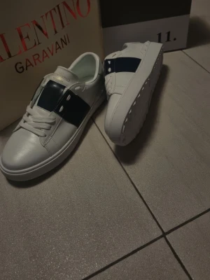 Valentino Garavani vita sneakers - Säljer ett par snygga Valentino Garavani sneakers i vitt skinn med en bred mörkblå rand och vita snören. Priset kan diskuteras! 
