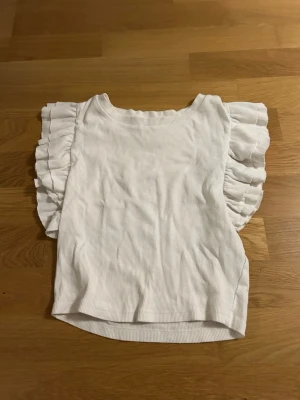 Vit ribbad topp med volangärmar från Zara - Säljer en vit ribbad topp från Zara i storlek M, kan även används som S. Toppen har breda, ärmlösa axlar med snygga volangdetaljer som ger en cool och trendig look. Materialet är mjukt och stretchigt, perfekt för att lyfta din outfit med lite extra stil. Säljer då den ej kommer till användning