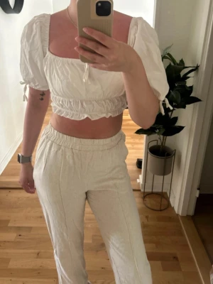 Vit croppad blus från NA-KD - Säljer en vit croppad blus från NA-KD med puffiga korta ärmar och volangdetaljer. Toppen har en fyrkantig ringning, knytband vid ärmslut och elastisk midja. Perfekt för sommarens alla tillfällen, superfin till både jeans och kjol.