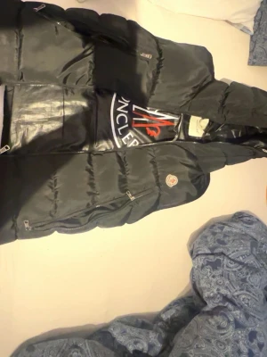 Svart dunväst från Moncler - Säljer en svart dunväst från Moncler med stor logga på insidan och detaljerade dragkedjor. Västen har quiltad design och Moncler-tryck längs insidan. Perfekt för dig som vill ha en stilren och sportig look.