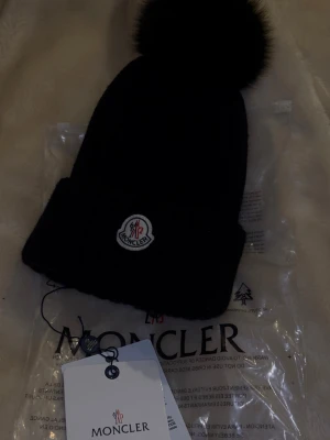 Svart stickad mössa med pälsboll - Säljer en svart stickad mössa med en fluffig pälsboll på toppen (äkta päls). Helt ny, enbart testat den. Den har qr kod som går att scanna