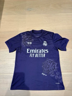 Lila Real Madrid x Y-3 fotbollströja - Säljer en limited edition Real Madrid x Y-3 fotbollströja i mörklila med blommigt mönster. Tröjan har tryck med 'Emirates Fly Better' och Real Madrid-logga på bröstet. Materialet är polyester och designen är modern med rund hals och korta ärmar.