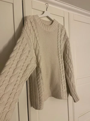 Beige stickad tröja med flätmönster - Säljer en beige stickad tröja med grovt flätmönster på ärmar och sidor. Tröjan har rund halsringning och långa ärmar med breda muddar. Perfekt för kyliga dagar och har en mysig, loose passform.