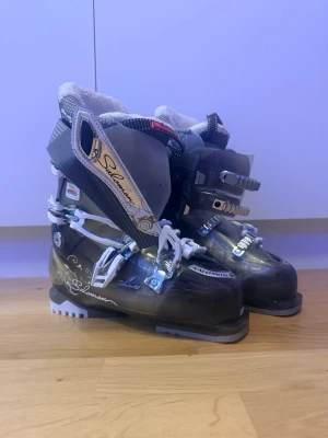 Salomon svarta skidpjäxor 25.5 - Säljer ett par svarta Salomon skidpjäxor med vita detaljer och metallspännen. Insidan är fodrad med mjukt, vitt material för extra komfort. Snyggt mönster på sidan och robust sula för bra grepp i backen. Perfekta för vinterns skidäventyr!