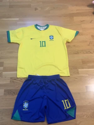 Brasilien Neymar Jr. matchställ - Säljer ett Brasilien matchställ med Neymar Jr. nummer 10. Tröjan är gul med gröna och blå detaljer, och har Brasil-logga samt Nike-logga på bröstet. Shortsen är blå med gula och gröna detaljer och har nummer 10 tryckt. Perfekt för dig som älskar fotboll och vill bära landslagets färger.