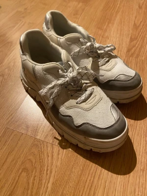 Vita sneakers från VOX Vegan Shoes - Säljer ett par chunky vita sneakers från VOX Vegan Shoes i storlek 39. Skorna har grå detaljer, rund tå och snörning med mönstrade vita skosnören. Tillverkade i syntetmaterial med mesh-paneler för extra ventilation. Perfekt för dig som vill ha veganska sneakers med cool stil.
